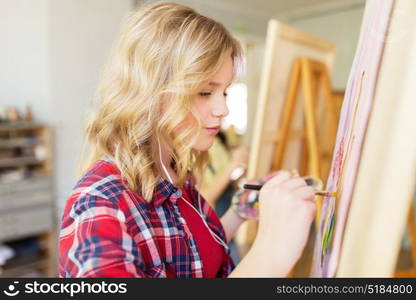 art school, creativity and people concept - student girl or artist with easel, earphones and paint brush painting at studio. student girl with easel painting at art school