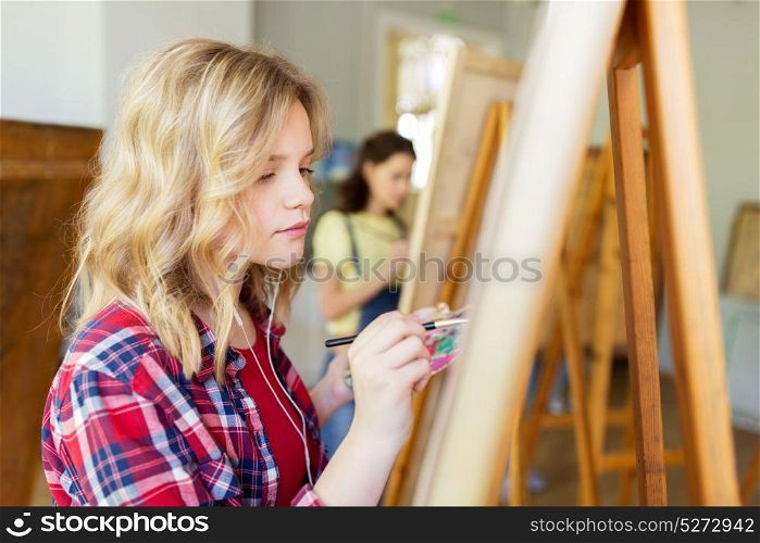 art school, creativity and people concept - student girl or artist with earphones, easel and paint brush painting at studio. student girl with easel painting at art school
