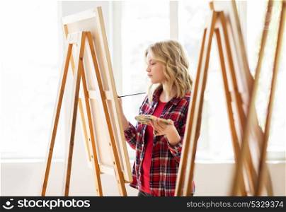 art school, creativity and people concept - student girl or artist with easel, palette and paint brush painting at studio. student girl with easel painting at art school