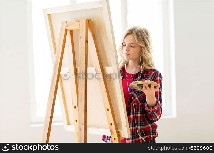 art school, creativity and people concept - student girl or artist with earphones, easel and palette painting at studio. student girl with easel painting at art school