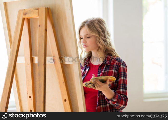 art school, creativity and people concept - student girl or artist with easel and palette painting at studio. student girl with easel painting at art school