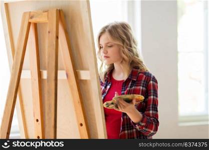 art school, creativity and people concept - student girl or artist with easel and palette painting at studio. student girl with easel painting at art school