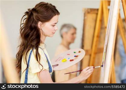art school, creativity and people concept - student girl or young woman artist with easel, palette and paint brush painting at studio. student girl with easel painting at art school