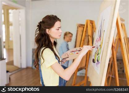 art school, creativity and people concept - student girl or young woman artist with easel, palette and paint brush painting still life picture at studio. student girl with easel painting at art school