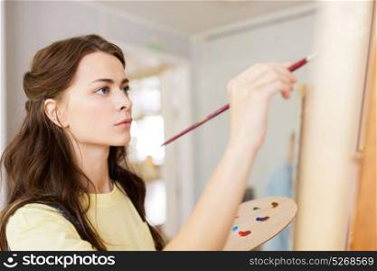 art school, creativity and people concept - student girl or young woman artist with easel, palette and paint brush painting at studio. student girl with easel painting at art school