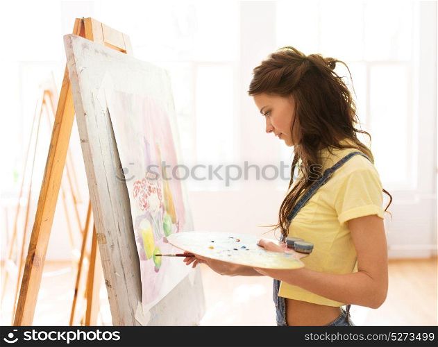 art school, creativity and people concept - student girl or young woman artist with easel, palette and paint brush painting still life picture at studio. student girl with easel painting at art school