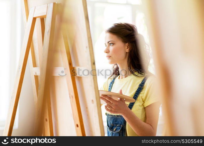 art school, creativity and people concept - student girl or young woman artist with easel and palette painting at studio. student girl with easel painting at art school