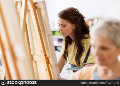art school, creativity and people concept - student girl or young woman artist with easel and palette painting at studio. student girl with easel painting at art school