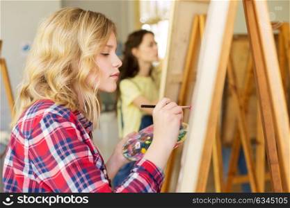 art school, creativity and people concept - teenage girl with easel, palette and brush painting at studio. girl with easel painting at art school studio