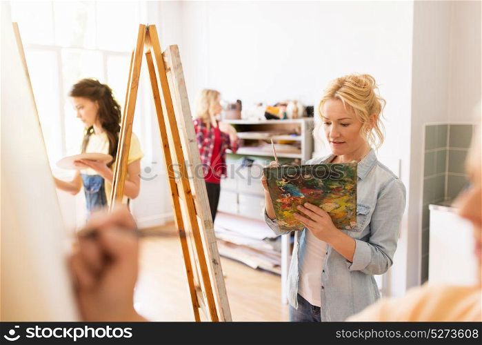 art school, creativity and people concept - woman artist with easel, paint brush and palette painting at studio. woman artist with easel painting at art school