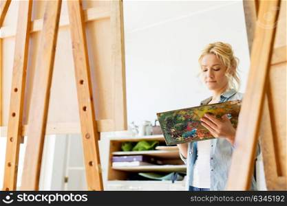art school, creativity and people concept - woman with easel and palette painting at studio. woman with easel painting at art school studio