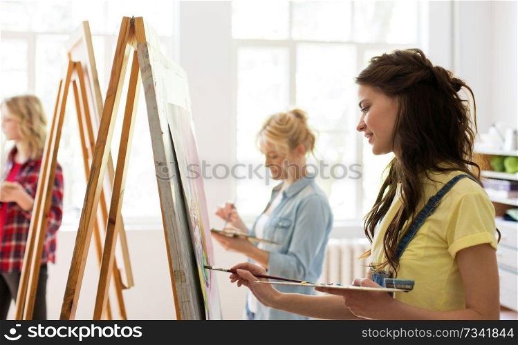 art school, creativity and people concept - woman with easel, palette and brush painting at studio. woman with easel painting at art school studio