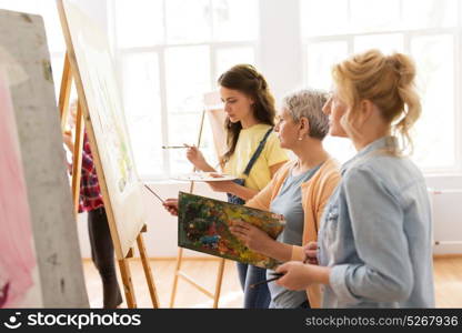 art school, creativity and people concept - women artists with easel, paint brushes and palettes painting at studio. women with easel and palettes at art school