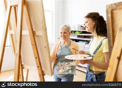 art school, creativity and people concept - women artists with easels, paint brushes and palettes painting at studio. women with easels and palettes at art school