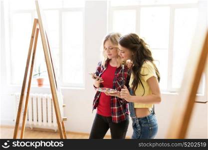 art school, technology and people concept - happy student girls or artists with smartphone, easel, palette and paint brush painting at studio. artists with smartphone at art studio or school
