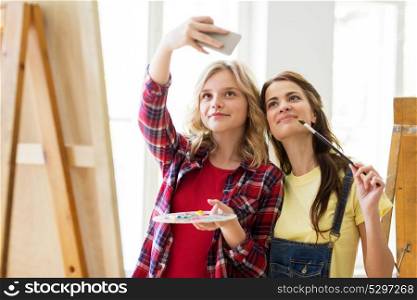 art school, technology and people concept - happy student girls or artists with smartphone, easel, palette and paint brush painting and taking selfie at studio. artist girls taking selfie at art studio or school