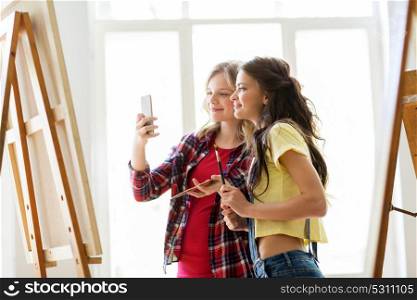 art school, technology and people concept - happy student girls or artists with smartphone, easel, palette and paint brush painting and taking selfie at studio. artist girls taking selfie at art studio or school