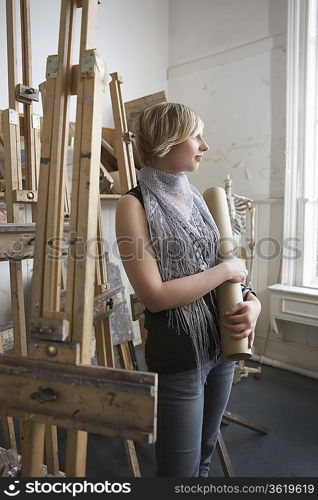 Art student standing, holding rolled-up artwork, in studio