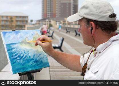 Artist standing on a pier and listening to music through earphones uses an angular brush to add highlights to a canvas mounted painting of the coast he is working on.