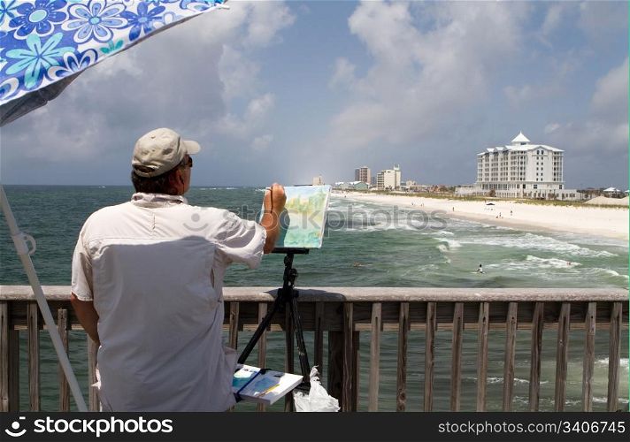 Artist uses a small brush to paint on canvas the view of the shoreline from a pier on Pensacola Beach, Florida.
