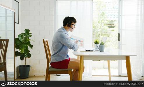 Asia businessman dressed in shirt and shorts use laptop talk to colleagues in video call while work from home at living room. Self-isolation, social distancing, quarantine for corona virus prevention.