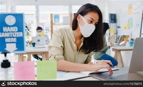Asia businesswoman entrepreneur wearing medical face mask for social distancing in new normal situation for virus prevention while using laptop back at work in office. Life and work after coronavirus.