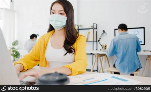 Asia businesswoman entrepreneur wearing medical face mask for social distancing in new normal situation for virus prevention while using laptop back at work in office. Lifestyle after corona virus.