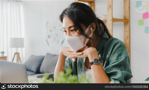 Asia businesswoman wearing medical face mask using laptop talk to colleagues about plan in video call while working from home at living room. Social distancing, quarantine for corona virus prevention.
