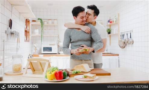 Asia gay couple using tablet and preparing the breakfast, sandwich vegetable on table in kitchen at home. Young LGBT men talking happy relax rest together spend romantic time at house in morning.