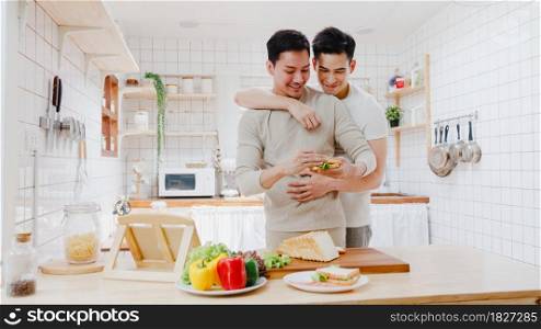 Asia gay couple using tablet and preparing the breakfast, sandwich vegetable on table in kitchen at home. Young LGBT men talking happy relax rest together spend romantic time at house in morning.