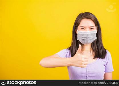 Asian adult woman wearing red shirt and face mask protective against coronavirus or COVID-19 virus or filter dust, air pollution her show finger thumb up for good sign, isolated yellow background