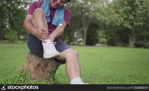 Asian aged male sprain his ankle during exercise Inside the park sitting on tree trunk with lawn on the background, ankle pain, joint ligament problem, painful face expression, risk of accident