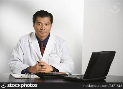 Asian American male doctor sitting at desk with charts and laptop computer looking at viewer.