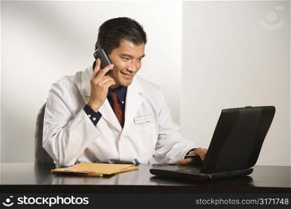 Asian American male doctor sitting at desk with charts and laptop computer talking on cellphone.