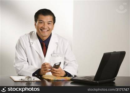 Asian American male doctor sitting at desk with charts and laptop computer using pda.