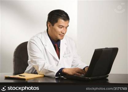 Asian American male doctor sitting at desk with charts typing on laptop computer.