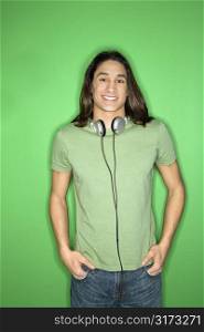 Asian-American teen boy smiling with headphones around neck and hands in pockets.