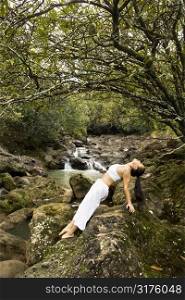 Asian American woman doing yoga balancing on boulder by creek in Maui, Hawaii.