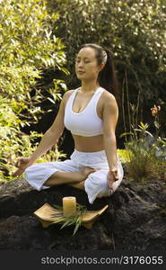 Asian American woman sitting on boulder in forest meditating with candle in Maui, Hawaii.