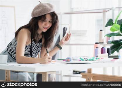 Asian attractive 30s woman fashion designer or dressmaker drawing sketch of new collection clothes on paper and smiling while working with tablet and tailoring equipments on desk at workplace studio