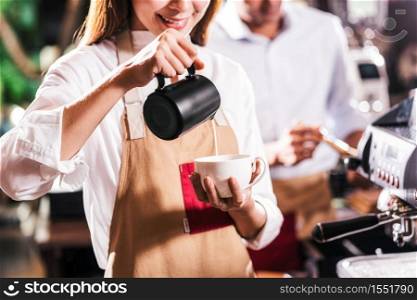 Asian Barista pouring milk to cup of coffee which is espresso with latte or cappuccino for customer order in coffee shop, Small business owner and startup in coffee shop and restaurant concept
