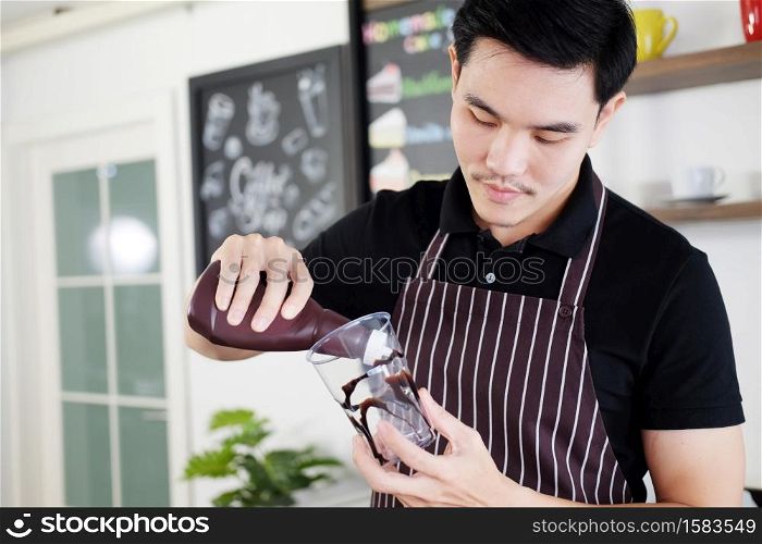 Asian barista young man pouring fresh Chocolate art in a plastic glass of iced coffee for according to order at counter bar in the morning at modern cafe and coffee shop.