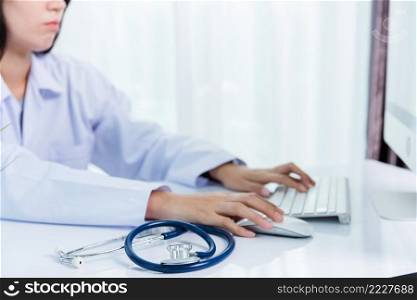 Asian beautiful female doctor smiling wearing doctor’s uniform with stethoscope typing on keyboard desktop computer looking to monitor on the desk at hospital office, Medical healthcare concept