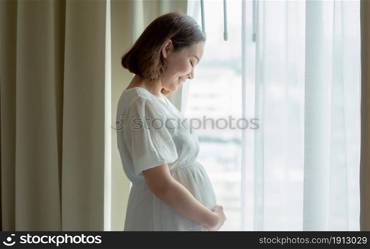 Asian beautiful pregnant woman wearing white dress and standing beside window in bedroom. Mother, Baby and Pregnancy Concept.