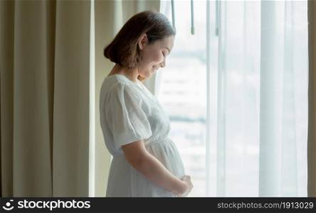 Asian beautiful pregnant woman wearing white dress and standing beside window in bedroom. Mother, Baby and Pregnancy Concept.