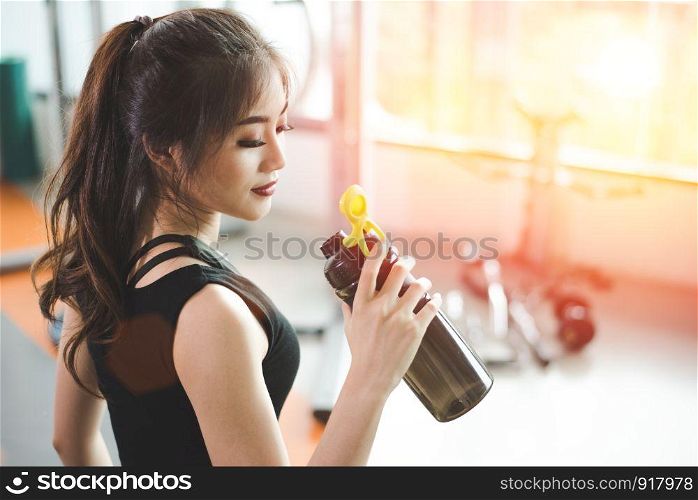 Asian beautiful woman drinking protein shake or drinking water in sport fitness training gym. Sports and people concept. Fitness and workout theme. Sun flare effect