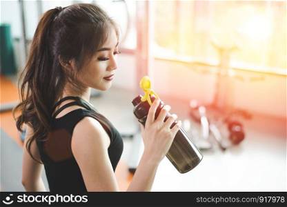 Asian beautiful woman drinking protein shake or drinking water in sport fitness training gym. Sports and people concept. Fitness and workout theme. Sun flare effect