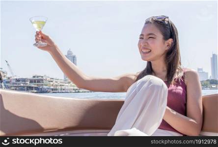 Asian beautiful woman drinking white wine and making toast while traveling and sitting on boat with background of beautiful building and landscape. Summer Travel and Lifestyle Concept.