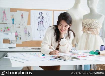 Asian beautiful woman fashion designer or dressmaker drawing sketch of new collection clothes on paper and smiling while working with tablet on desk at workplace studio