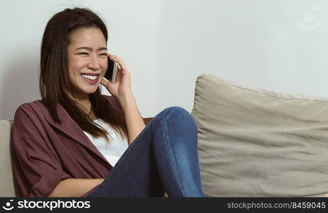 Asian beautiful woman smiling and talking on her mobile phone while sitting on sofa in living room at home. Lifestyle and Technology Concept.